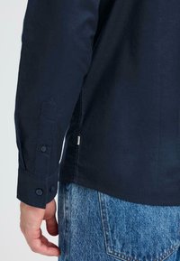 Camisa de manga larga azul marino con puños abotonados, hecha de algodón, con una etiqueta lateral y un dobladillo redondeado, combinada con jeans de mezclilla claros.