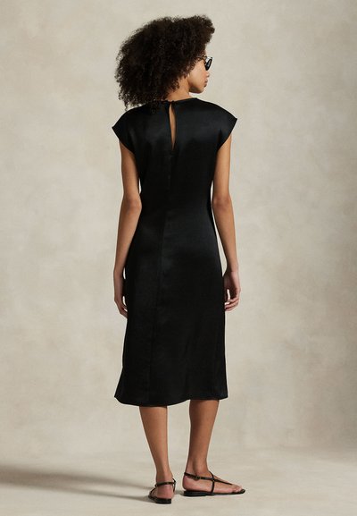 Vestido de cetim preto com mangas curtas em formato de capa, costas com abertura em forma de gota e comprimento até ao joelho. Modelo combinado com sandálias pretas minimalistas.