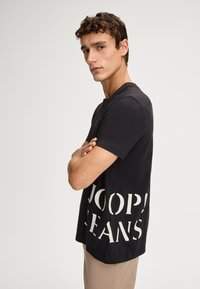 Czarny bawełniany t-shirt z krótkim rękawem, z dużym białym napisem "JOOP! JEANS" nadrukowanym z boku. Prosty krój, minimalistyczny design.