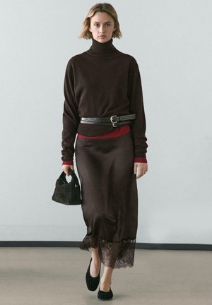 Maglione marrone a collo alto con dettagli rossi, abbinato a una gonna marrone di raso con orlo in pizzo. La modella tiene una borsa nera testurizzata. Scarpe nere.