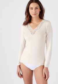 Maglia a maniche lunghe color crema con scollatura a V profonda e bordo in pizzo. Tessuto morbido con design aderente. Indossata con lingerie bianca, evidenziando una silhouette morbida.
