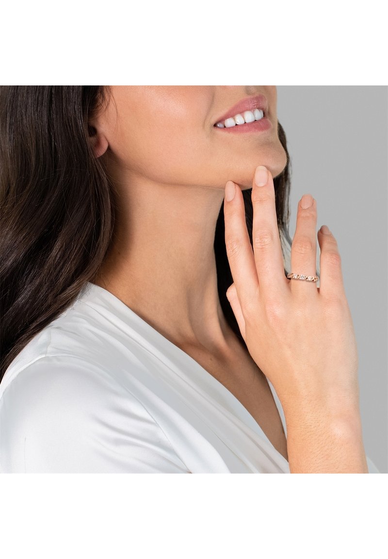 Guldtonet ring med intrikat sammenflettet design. Modellens hånd hviler på hendes kæbe, hvilket fremhæver ringens detaljer. Blød, neutral hudtone.