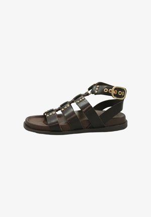 Sandalo gladiatore in pelle marrone con fibbia dorata e borchie metalliche, suola piatta e più cinturini sul piede e sulla caviglia.