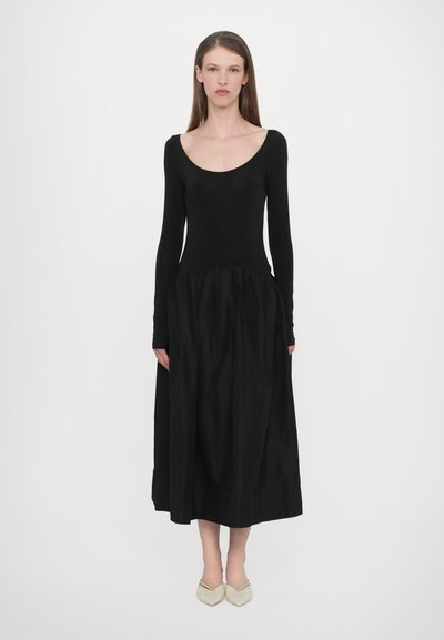 Sort langærmet kjole med en rund halsudskæring og rynket skirt, lavet af et glat stof. Sammen med lyse, spidse sko.