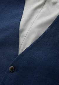 Gros plan sur le revers d'un blazer bleu marine avec un unique bouton foncé, révélant une chemise blanche à motif en dessous.