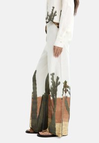 Spodnie lniane o szerokich nogawkach z nadrukiem przedstawiającym pustynny krajobraz z kaktusami i palmami w stonowanych odcieniach zieleni, brązu i beżu. W zestawie czarne płaskie sandały.