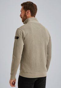 Sudadera cremosa con cremallera y alto cuello, de textura acanalada, con un parche negro en el hombro izquierdo. Se ven pantalones negros.