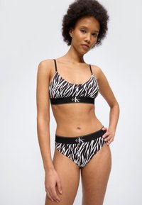 Γυναίκα που φοράει μαύρο και λευκό σουτιέν με σχέδιο ζέβρας Calvin Klein και ταιριαστό εσώρουχο, ποζάροντας με το ένα χέρι στη μέση της μπροστά από έναν απλό φόντο.