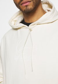 Sudadera crema hecha de un tejido suave con un bolsillo tipo canguro. Cuenta con una capucha adjustable y un estampado de logo blanco en el frente.