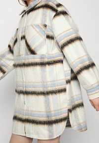 Chaqueta de camisa de cuadros en material suave y peludo; base crema con rayas azul claro, gris y negro; dos bolsillos frontales; diseño de botones.