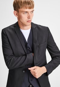 Black blazer with a tailored design, features a single breast pocket, notched lapels, and textured fabric. Worn over a dark vest and white shirt.
