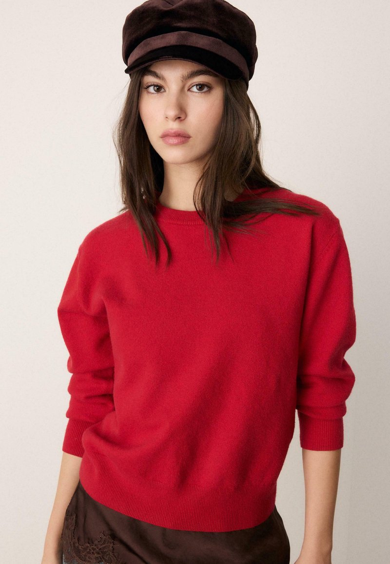Pull tricot rouge à manches longues et col rond. Texture douce et ourlet côtelé. Porté avec une casquette marron foncé et superposé sous une jupe sombre.