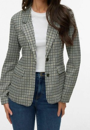 Femme portant un blazer pied-de-poule noir et blanc par-dessus un haut blanc et un jean bleu, avec de longs cheveux bruns partiellement visibles.
