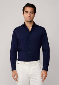 Granatowa koszula z długim rękawem, z kołnierzykiem na guziki, o dopasowanym kroju i delikatnym połysku, zestawiona z białymi spodniami i ciemnymi guzikami.