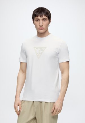 Homme portant un t-shirt blanc à manches courtes avec un logo Guess en relief subtil, associé à un pantalon beige, debout sur un fond uni.