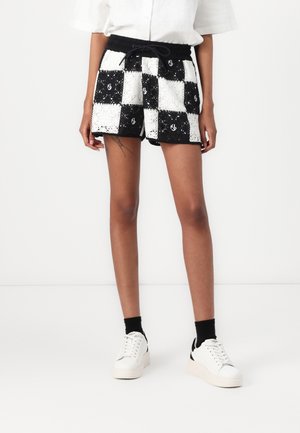 Persoon draagt zwart-wit geruite gehaakte shorts, witte sneakers met zwarte sokken en een wit T-shirt met korte mouwen, staand tegen een witte achtergrond.