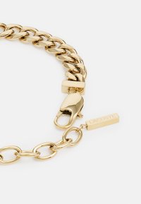 Goldene Kettenarmband mit ineinandergreifenden Gliedern, einem Karabinerhaken und einem rechteckigen Anhänger mit dem gravierten Markennamen "Lacoste".