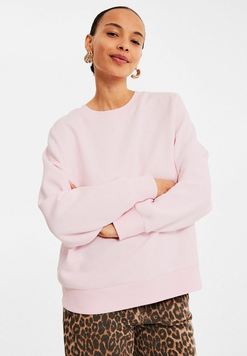 Helles pinkes Sweatshirt mit Rundhalsausschnitt, weiten Ärmeln und gerippten Bündchen, kombiniert mit braunen Leopardmuster-Hosen. Glatte Stofftextur.