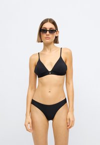 Σετ μαύρου μπικίνι με τριγωνικό σουτιέν με λεπτές τιράντες και χρυσή λεπτομέρεια, συνδυασμένο με αντίστοιχα κάτω μέρη χαμηλής μέσης. Ομαλή υφή.