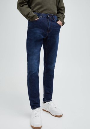 Homme portant un jean slim bleu marine, un sweat-shirt vert olive et des baskets blanches, debout les mains dans les poches devant un fond uni.