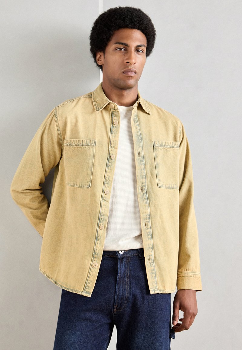 Camasa de denim galbenă cu două buzunare la piept, închidere cu nasturi și croială lejeră, purtată peste un tricou alb cu dungi și blugi albaștri.