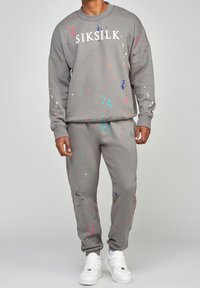 Conjunto de sudadera y pantalones de chándal grises con manchas de pintura multicolores. Presenta un escote redondo y un logo blanco. Zapatillas deportivas blancas.