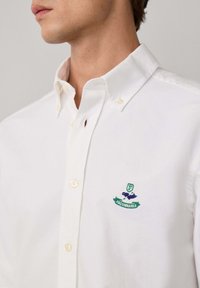 Primer plano de una persona que lleva una camisa blanca de botones con un logo bordado en verde y azul de Façonnable en el pecho.