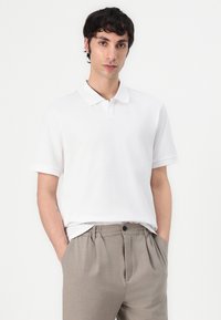 Vit polo med kort ärm, platt krage och diskret logotypdetalj, i kombination med ljusbeige byxor med elastisk midja och fickor.