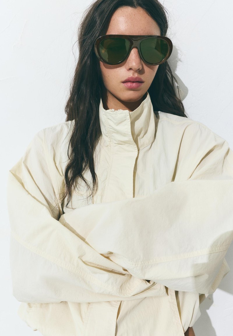 Femme aux longs cheveux foncés portant de grandes lunettes de soleil foncées et une veste crème à col haut, debout sur un fond blanc.