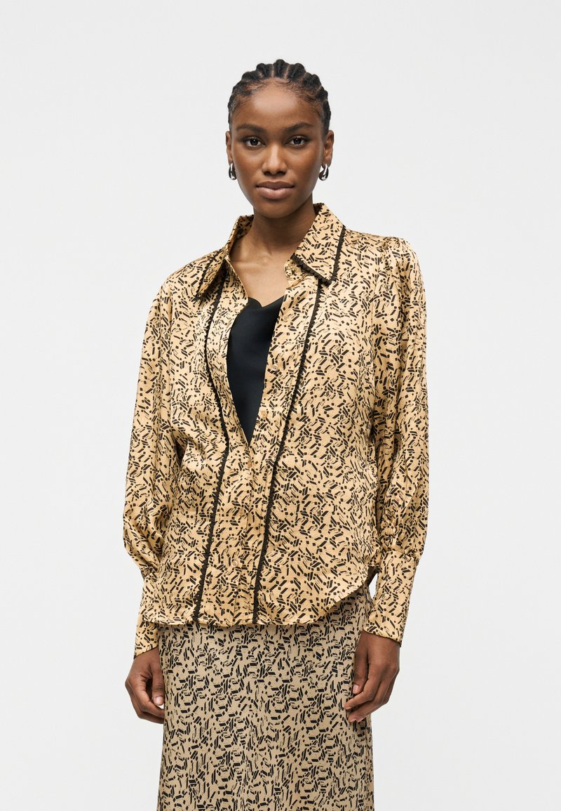 Γυναίκα με πλεξούδες που φοράει μπεζ σακάκι με μοτίβο και ταιριαστή φούστα πάνω από μαύρη μπλούζα, κοιτάζοντας μπροστά με ουδέτερη έκφραση.