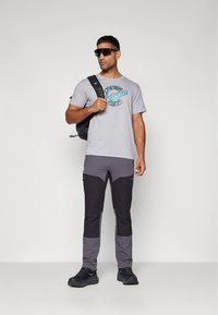 Hall T-särk türkiissinise graafikaga, mustade ja hallide värviplokiga pükstega ning musta seljakotiga. Modell kannab päikeseprille ja tumedaid kingi.