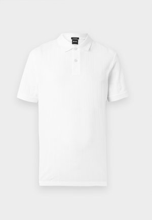 Hvid polo med korte ærmer, to-knaps stolpelukning og en subtil lodret ribstruktur, lagt fladt på en lys baggrund.