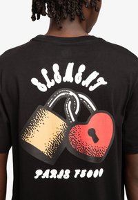 Zwarte t-shirt met een grafisch ontwerp van een hangslot en een hart, met textuurdetails en de tekst "Paris T5000" eronder.