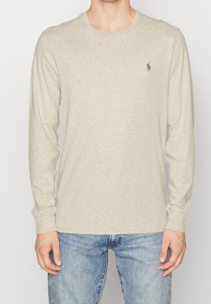 Man draagt een effen beige shirt met lange mouwen en een ronde hals, met een klein logo van een paard en ruiter op de linkerborst, gecombineerd met lichtblauwe spijkerbroek.