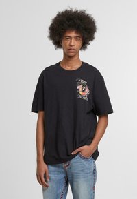 Camiseta de algodón negra con un gráfico frontal que presenta el texto "True Religion" en una tipografía colorida. Corte de manga corta y un diseño de ajuste relajado.