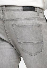 Un jean en denim gris présente une poche arrière avec un design subtil, cousue avec un fil assorti. La taille comporte une étiquette noire marquée "INDICODE."