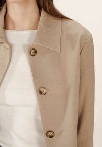 Primo piano di una giacca in suede beige con tre bottoni marmorizzati sopra una maglietta bianca a coste indossata da una persona.