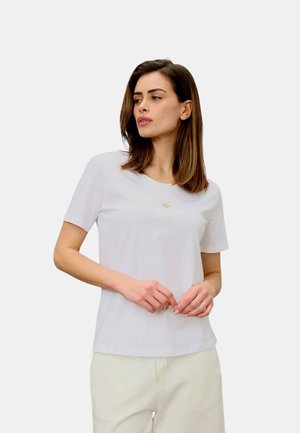 Donna con capelli castani fino alle spalle indossa una maglietta bianca semplice a maniche corte e pantaloni chiari, guarda leggermente di lato con le mani giunte.