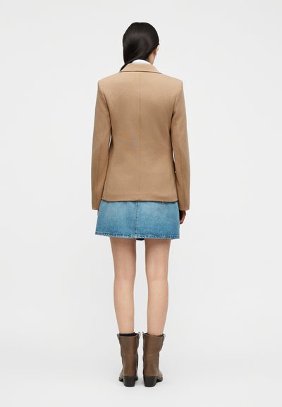 Γυναίκα με την πλάτη, φορώντας ένα σακάκι καμήλας, ένα μπλε τζιν μίνι φούστα και καφέ μποτάκια αστραγάλου σε απλό λευκό φόντο.