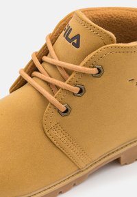 Stivaletto alla caviglia in pelle tan con occhielli metallici esagonali, lacci beige e superficie texturizzata con suola in gomma marrone. Logo Fila presente.