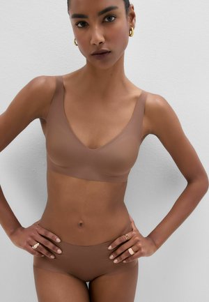 Kvinde iført sømløs brun bh og matchende undertøj, stående med hænderne på hofterne mod en ensfarvet lys væg.