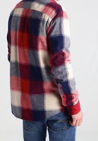 Karierte Flanellhemd in Rot, Blau und Creme mit langen Ärmeln, einzelnen Knopfmanschetten und abgerundetem Saum. Weicher Stoff, sichtbare Nahtdetails.