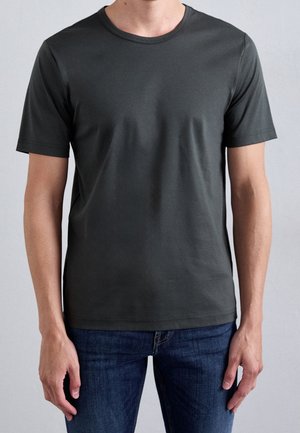Mężczyzna w prostym ciemnoszarym t-shircie z krótkim rękawem i niebieskich dżinsach, stojący na jasnoszarym tle.