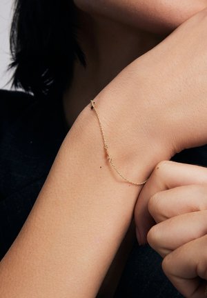 Złoty łańcuszek bransoletka o delikatnym wzorze z pojedynczym akcentem, noszona na nadgarstku w zestawieniu z neutralnym odcieniem skóry.