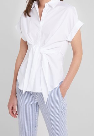 Vrouw draagt een witte blouse met korte mouwen, gestrikt in de taille, en blauw-wit gestreepte broek met verticale strepen, met één hand in de zak.