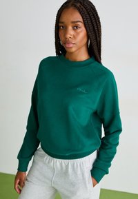 Sudadera verde con cuello redondo y puños de canalé, llevada sobre pantalones de chándal grises. Presenta un discreto logo bordado en el pecho.