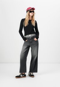 Vrouw met lang blond haar, draagt een zwart shirt met lange mouwen, wijde vervaagde zwarte jeans, zwarte sneakers, een rode pet en zwarte zonnebril, staand.