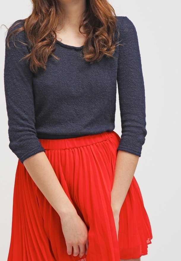 Femme aux cheveux bruns ondulés portant un haut à manches longues bleu marine foncé et une jupe plissée rouge vif, les mains doucement jointes devant.