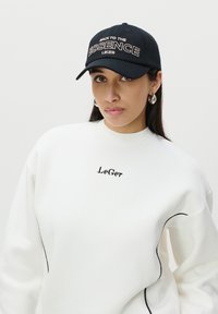 Sweatshirt blanc avec des coutures noires et le logo "LeGer". Casquette noire avec un texte brodé "BACK TO THE ESSENCE" en couleur claire. Boucles d'oreilles rondes.