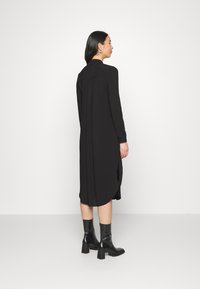 Vestido preto de manga longa com um corte descontraído, fendas laterais e colarinho. Usado com botas de cano curto pretas e brilhantes. Textura de tecido suave.